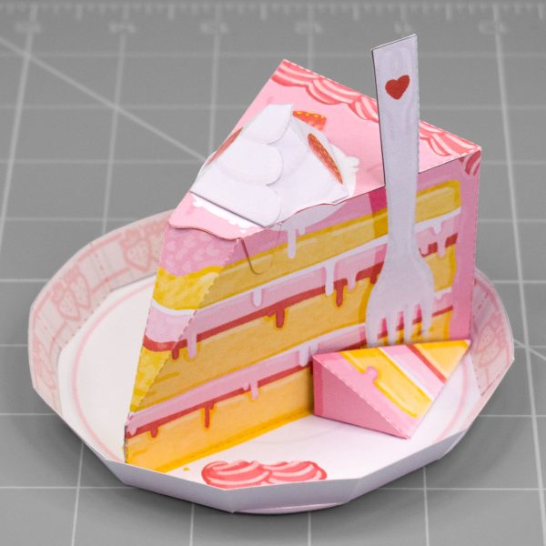 A photo of a papercraft depicting a slice of strawberry cake on a decorative plate. The papertoy has a fork sticking out of the side and has whipped cream on the top.