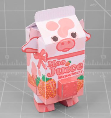 A photo of a papercraft depicting a carton of strawberry milk, with a cow's features, such as ears, udders and a mouth.